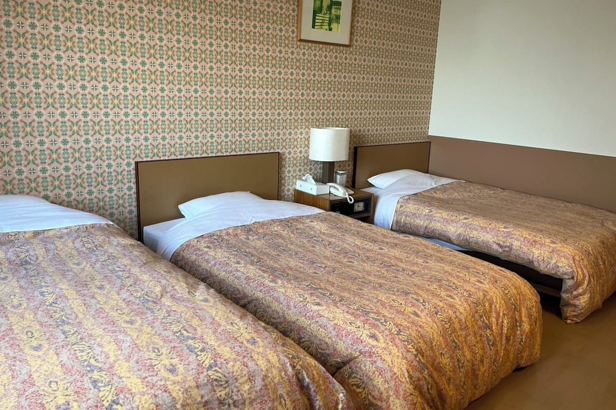 Quarto de hotel com três camas, cada uma com roupa de cama estampada, mesa de cabeceira com abajur e quadro na parede.