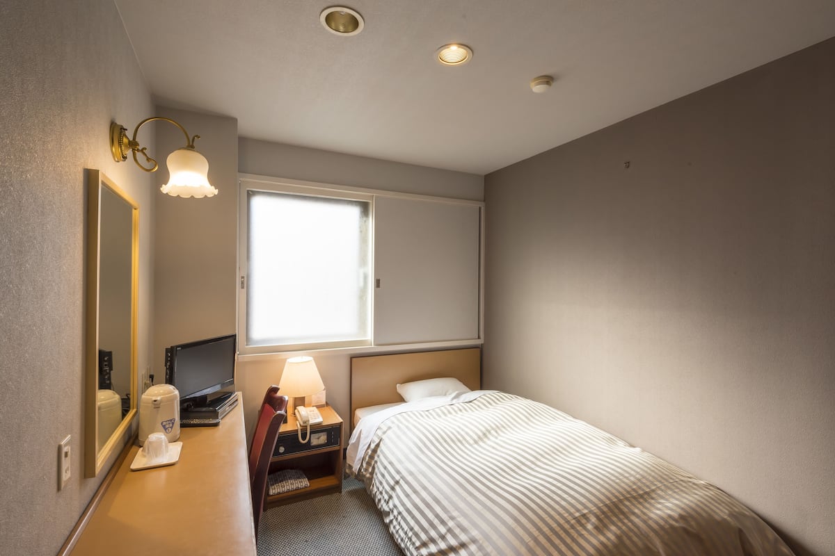 Quarto de hotel com cama, escrivaninha, computador, telefone, luminária, espelho e janela.