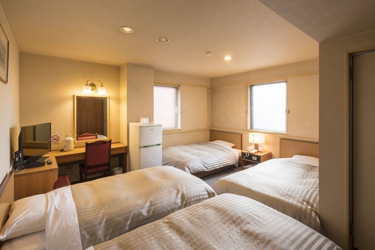Quarto de hotel com duas camas, uma escrivaninha com computador, um frigorífico e uma cadeira.