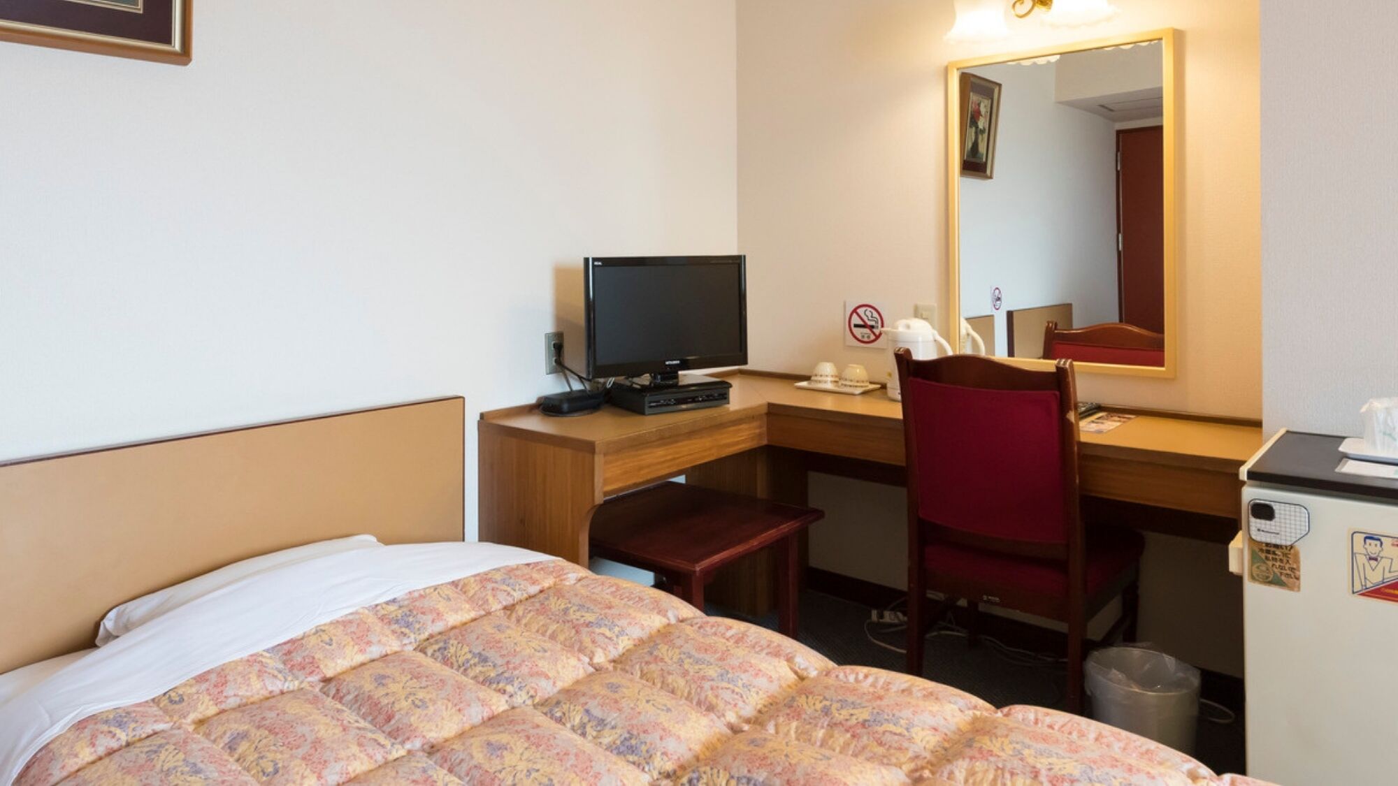 Habitación de hotel con cama, escritorio con computadora, silla, espejo y refrigerador pequeño.