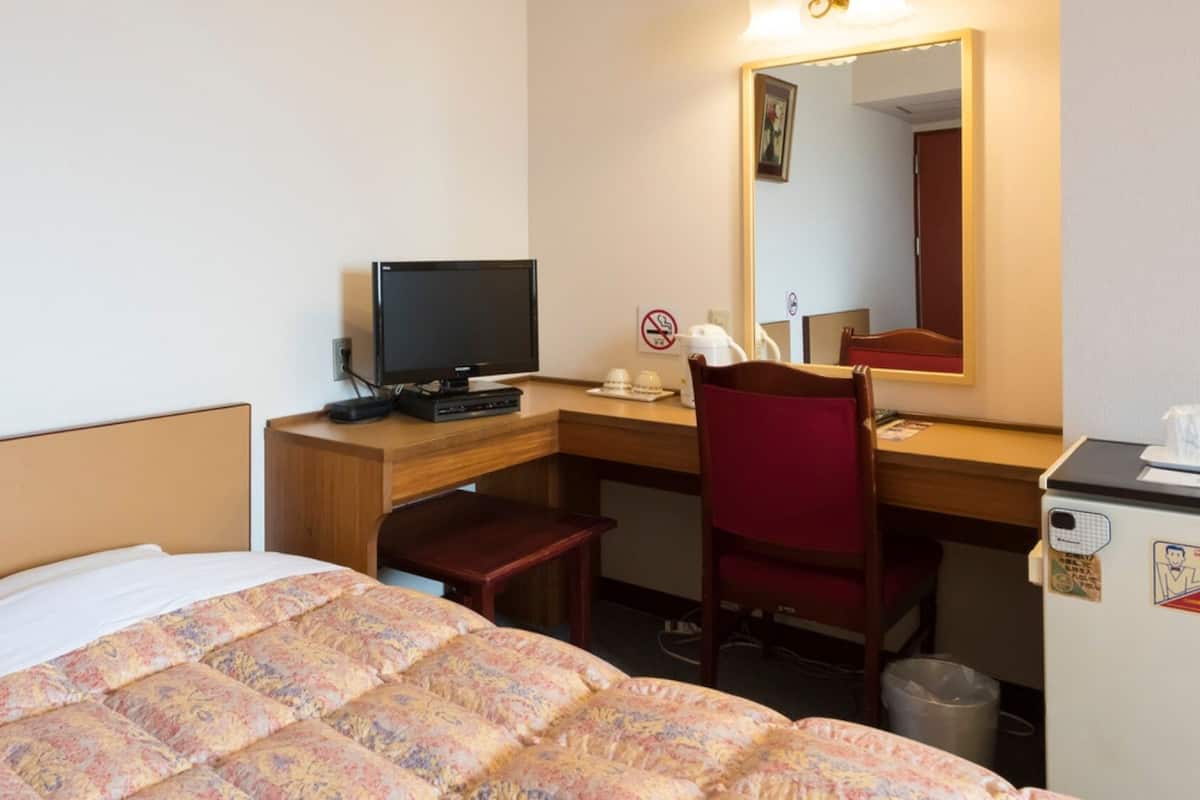 Quarto de hotel com uma cama, uma escrivaninha com computador, uma cadeira, um espelho e um pequeno frigorífico.