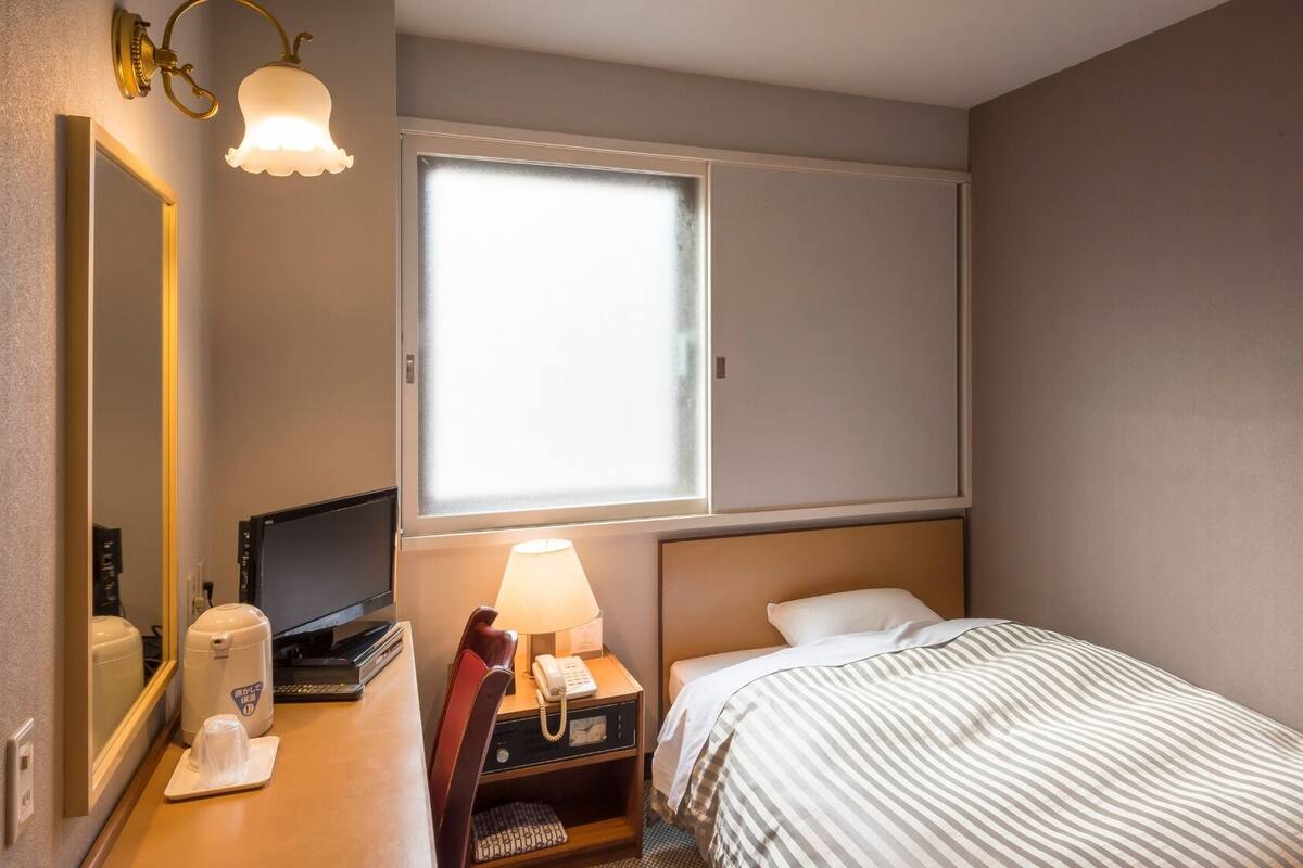 Quarto de hotel com cama, secretária com telefone, luminária, espelho e janela com persianas.