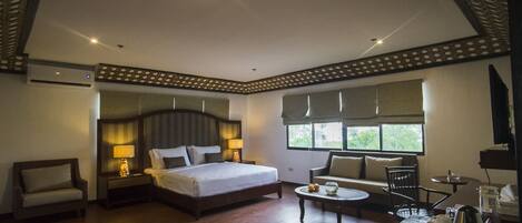 A spacious hotel room with a large bed, a sitting area with a coffee table, and a view of the outdoors through the windows.