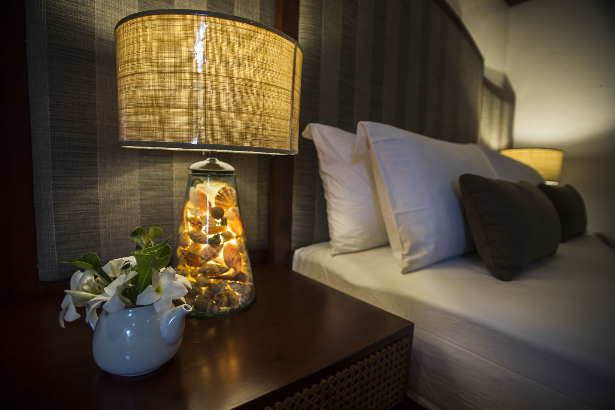 Un lit bien fait, avec une literie blanche et grise, une lampe de chevet dotée d’un abat-jour en osier, et un bocal en verre contenant des fleurs blanches et des pierres décoratives.