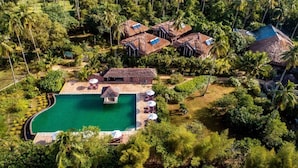 Vista aérea de um resort com piscina central rodeada por edifícios com telhados de palha e vegetação exuberante.