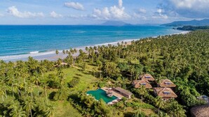 Um resort tropical com piscina, bangalôs de palha e uma praia.