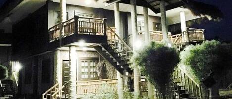 A two-story building with a balcony on the upper floor, a staircase leading to the upper level, and a covered outdoor area with a thatched roof.