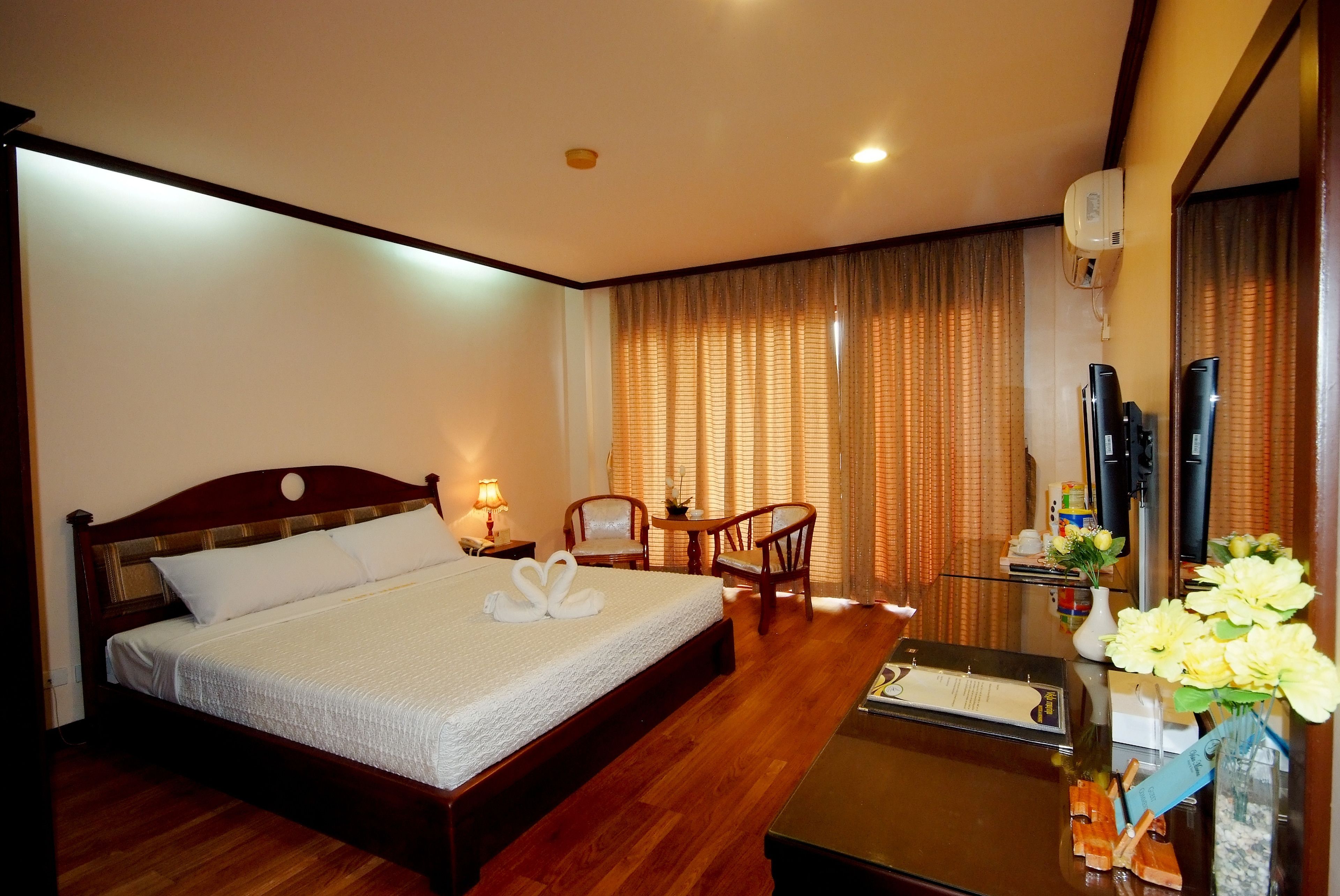 Une chambre d’hôtel avec un lit en bois, deux chaises, une petite table, un téléviseur et un vase avec des fleurs.
