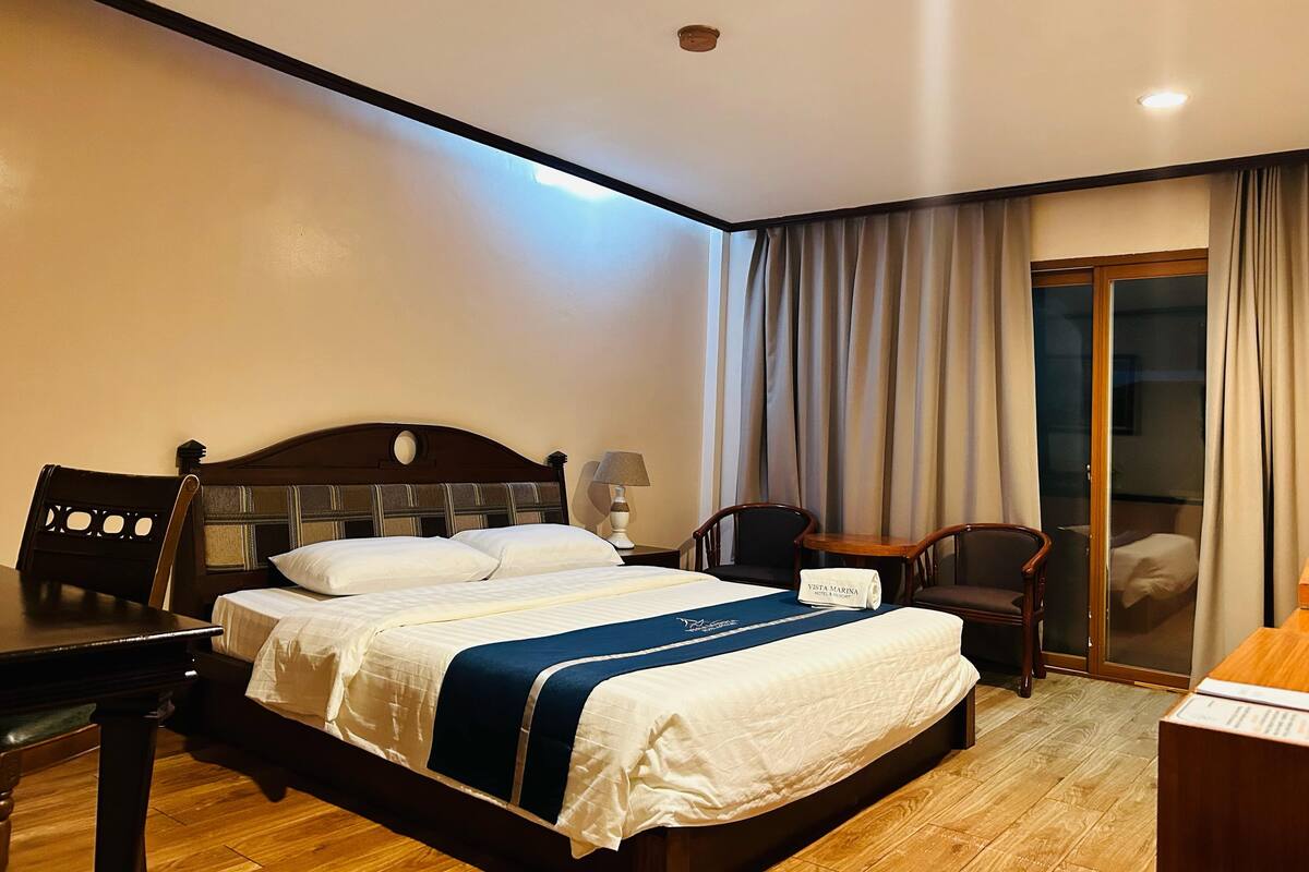 A hotel room with a bed, a desk, a chair, a nightstand, and a sliding glass door to a balcony.