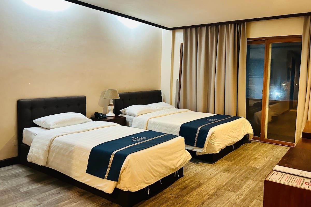 A hotel room with two beds, a nightstand with a lamp, and a sliding glass door leading to a balcony.