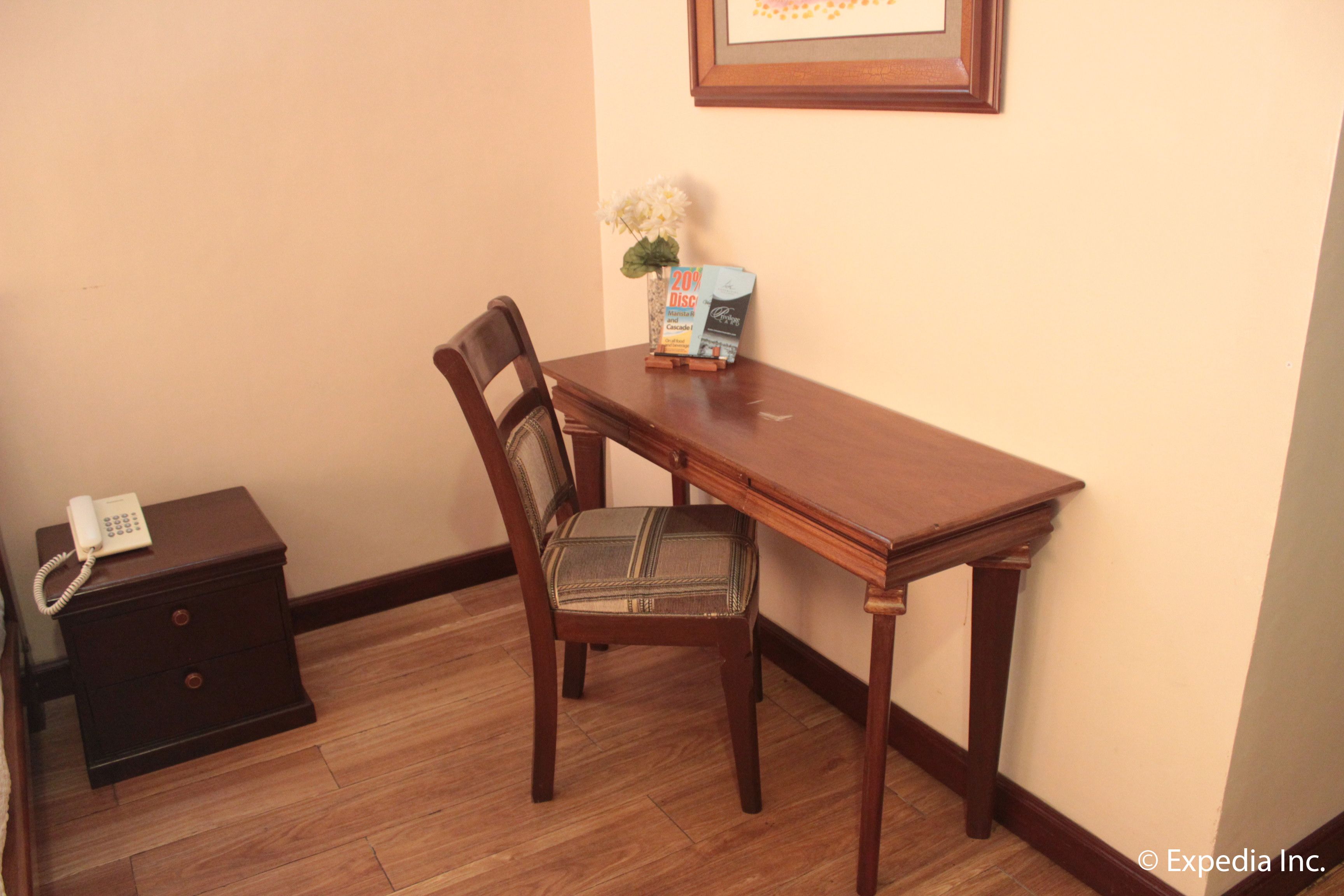 Une table de salle à manger en bois, accompagnée d’une chaise, une petite table de chevet en bois avec un téléphone, et un tableau encadré accroché au mur.