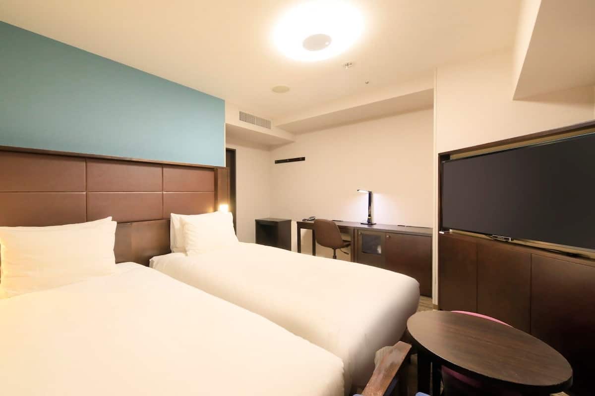 Quarto de hotel com duas camas, uma escrivaninha, uma cadeira, uma televisão e uma mesa redonda pequena.