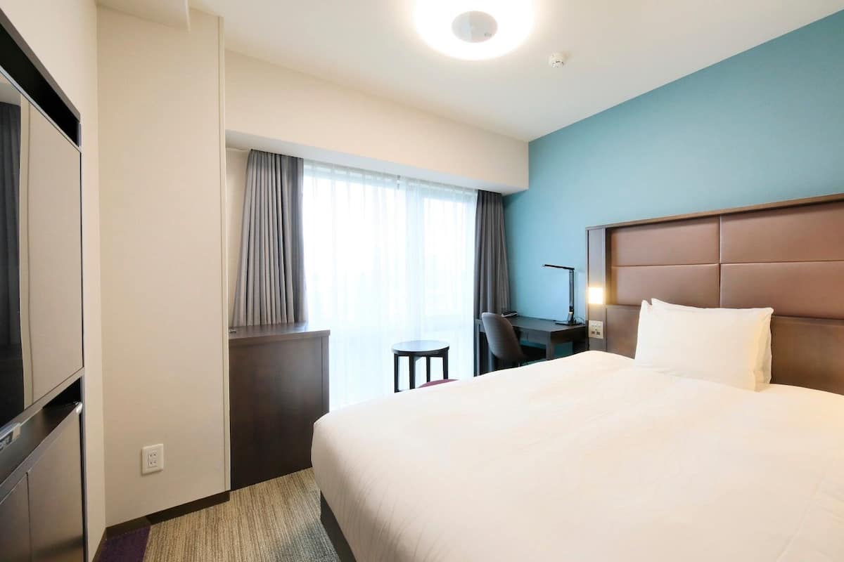 Quarto de hotel com uma cama grande, uma escrivaninha com cadeira, televisão e janela com cortinas.