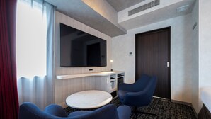 A hotel room with a flat-screen TV, a round table, two blue chairs, a wooden cabinet, and a dark door. - Richmond Hotel Namba Daikokucho (Osaka)