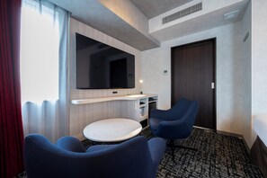Habitación de hotel con televisor de pantalla plana, una mesa redonda, dos sillas azules, un armario de madera y una puerta oscura.