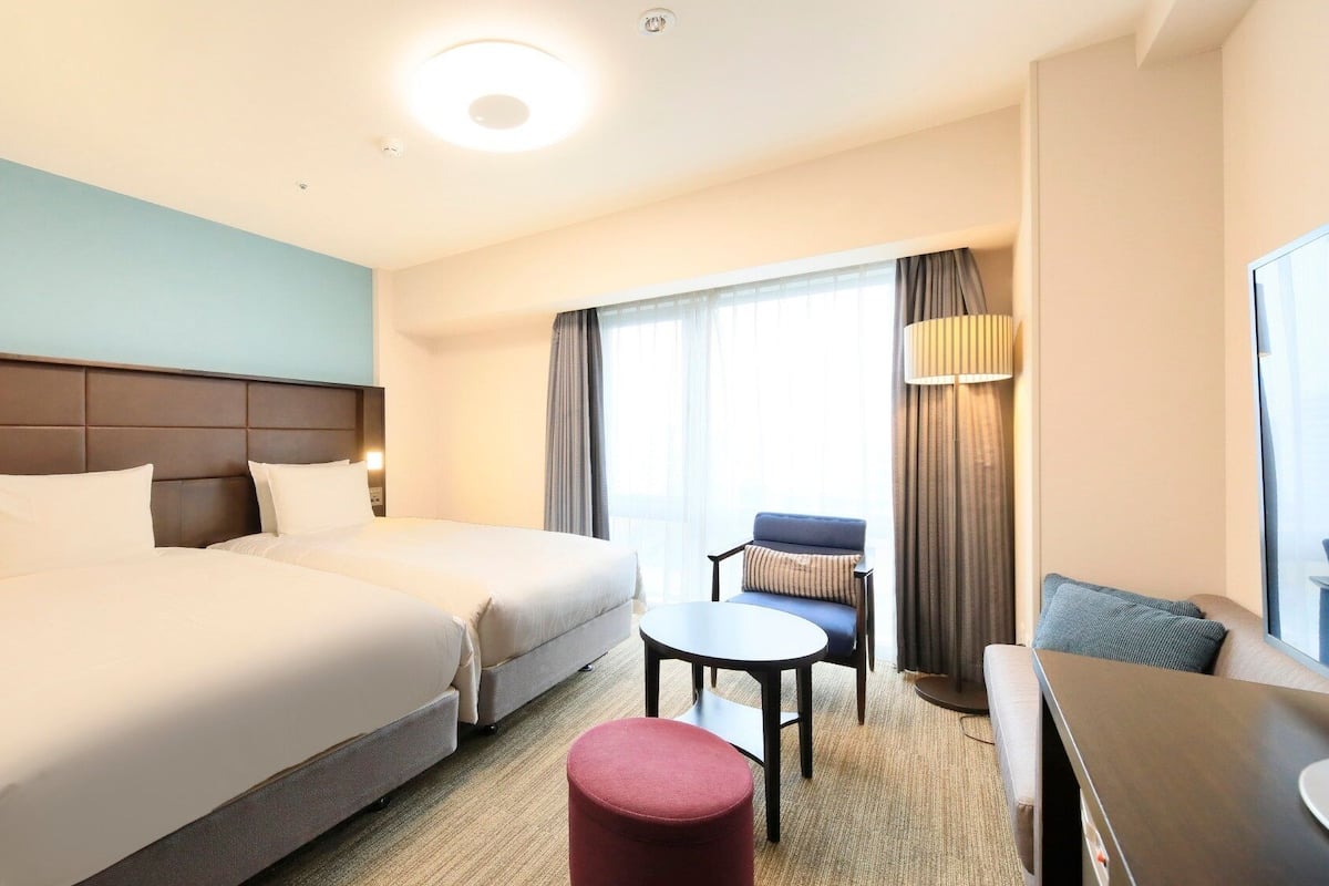 Quarto de hotel com duas camas, uma escrivaninha, uma cadeira, uma mesinha e uma janela com cortinas.