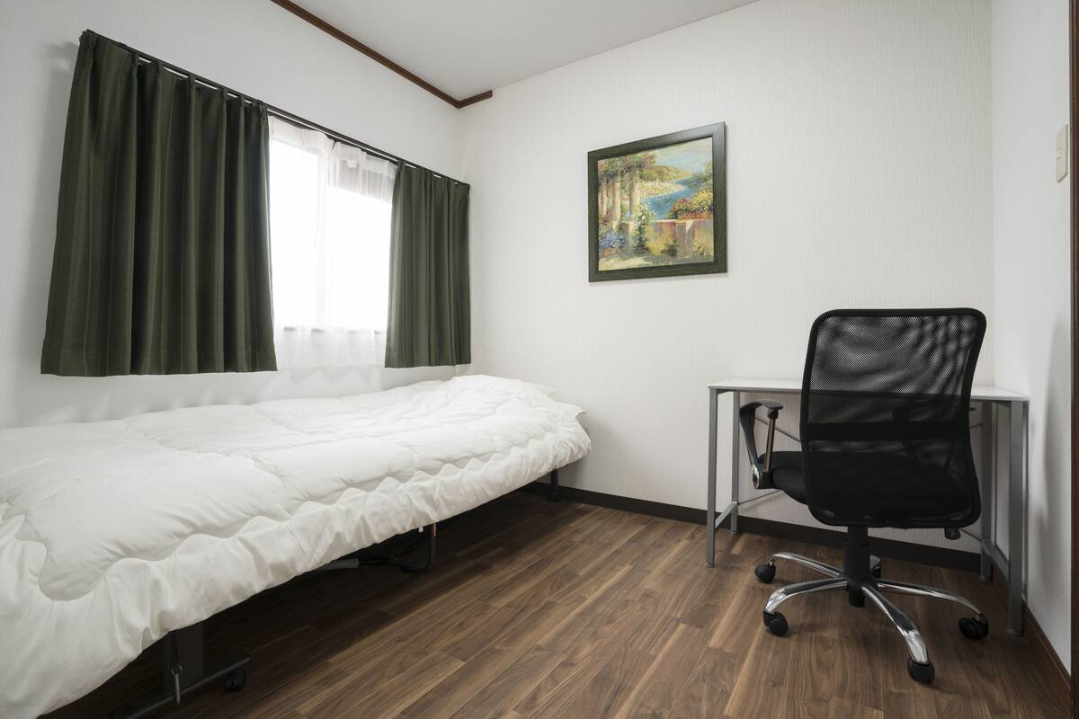 Una cama individual con edredón blanco, una silla de escritorio negra y un cuadro en la pared.