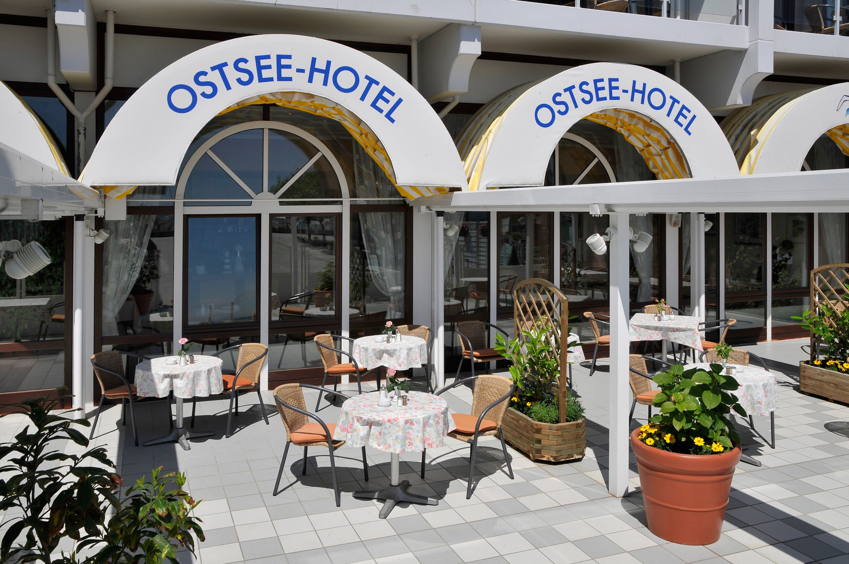 Ein Außenbereich mit Tischen und Stühlen, großen Kübelpflanzen und freiem Blick auf den Eingang des Hotels.