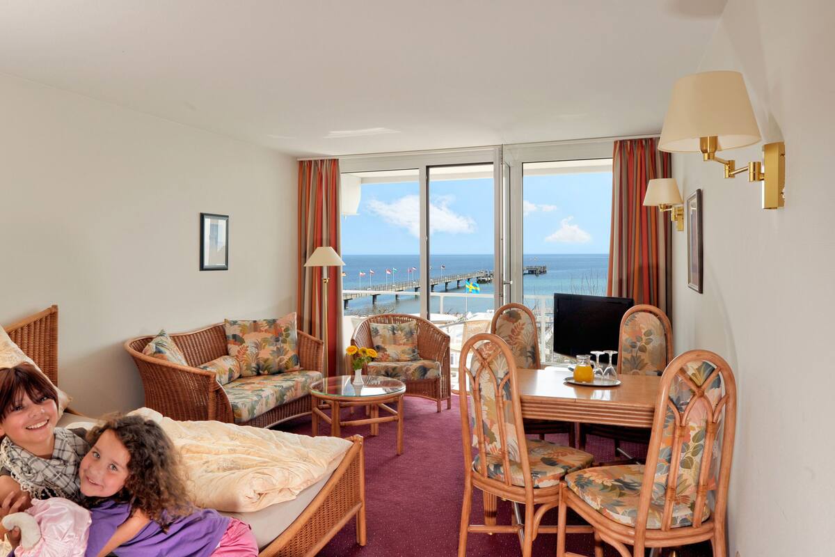 Ein Hotelzimmer mit großem Fenster, Korbmöbeln, einem Esstisch und einem Fernseher.