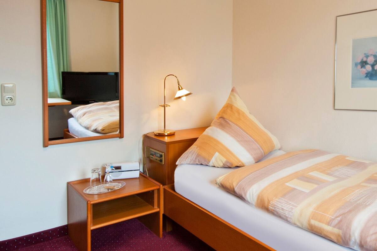 Ein Hotelzimmer mit einem Bett, einem Nachttisch, einer Lampe, einem Spiegel, einem Fernseher und einem gerahmten Bild an der Wand.