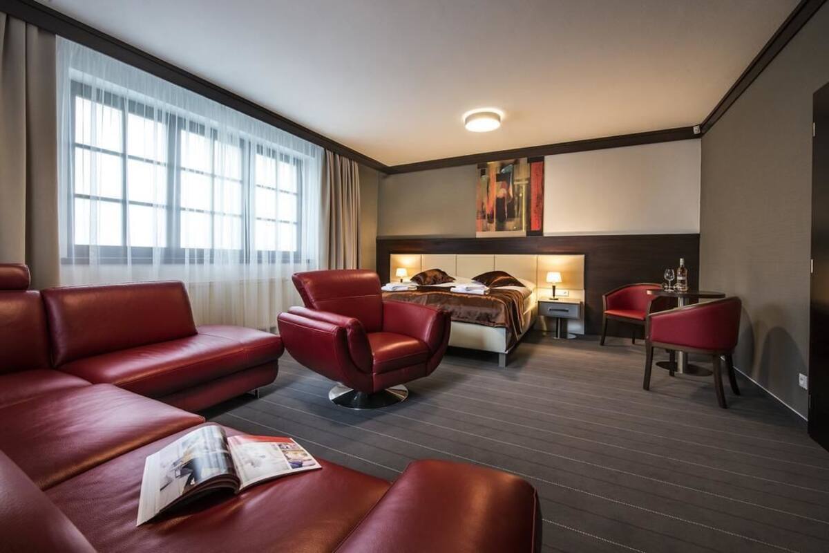 A hotel room with a large window, a bed, a desk with a chair, and a red leather sofa.