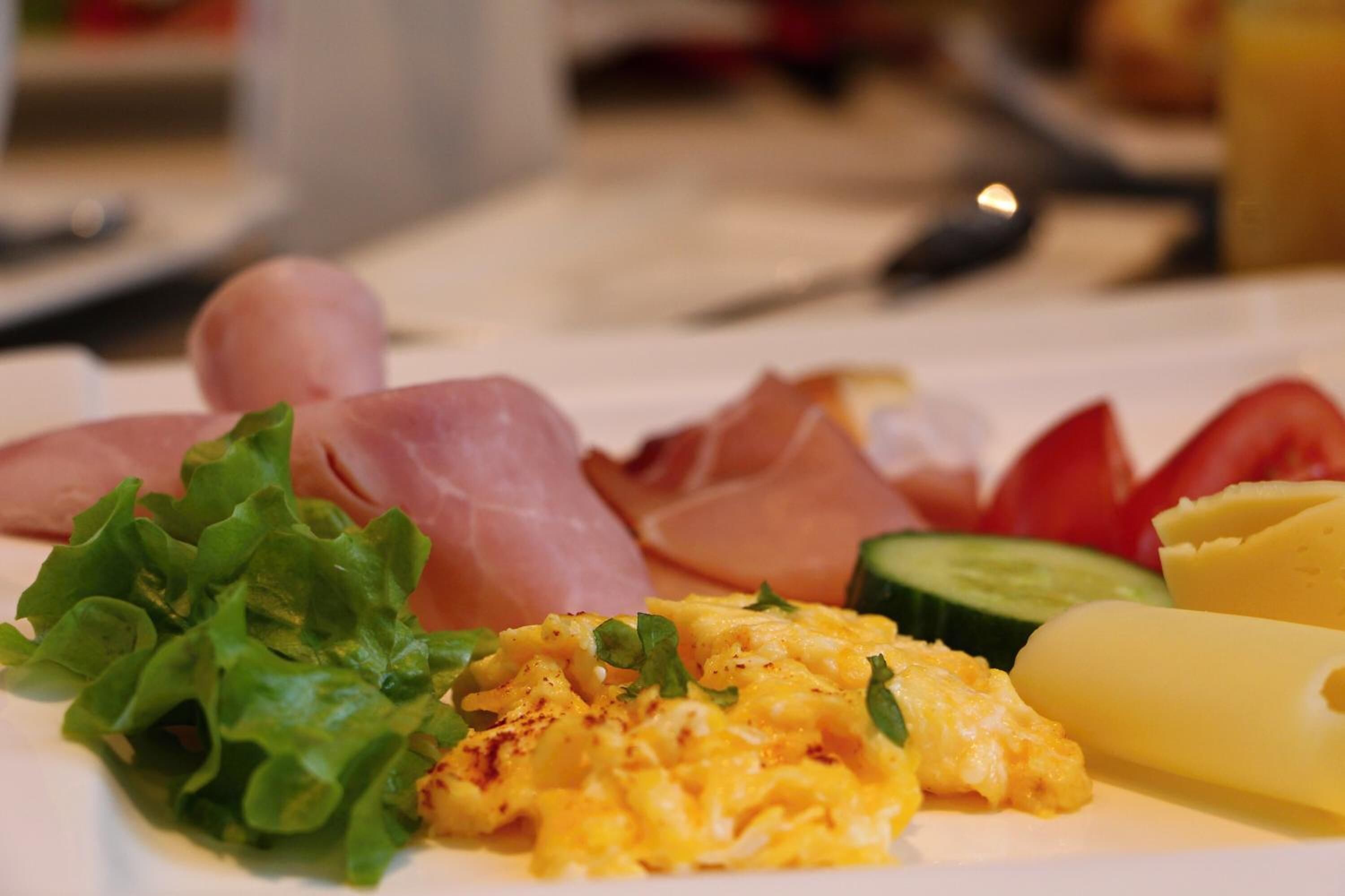 A plate with scrambled eggs, ham, prosciutto, cucumber, lettuce, and a slice of cheese.