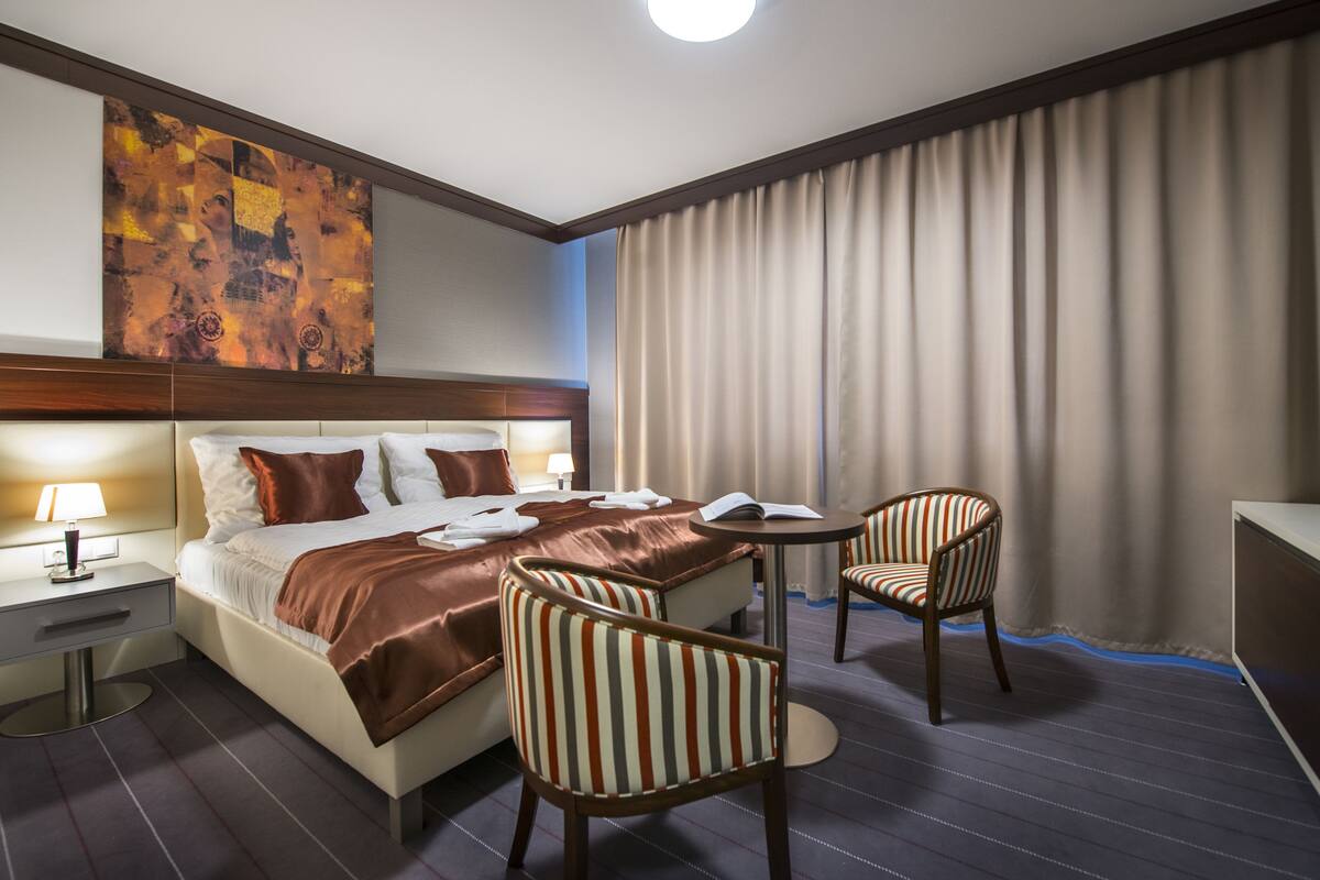 A hotel room with a large bed, two striped chairs, a small table, and a large abstract painting on the wall.