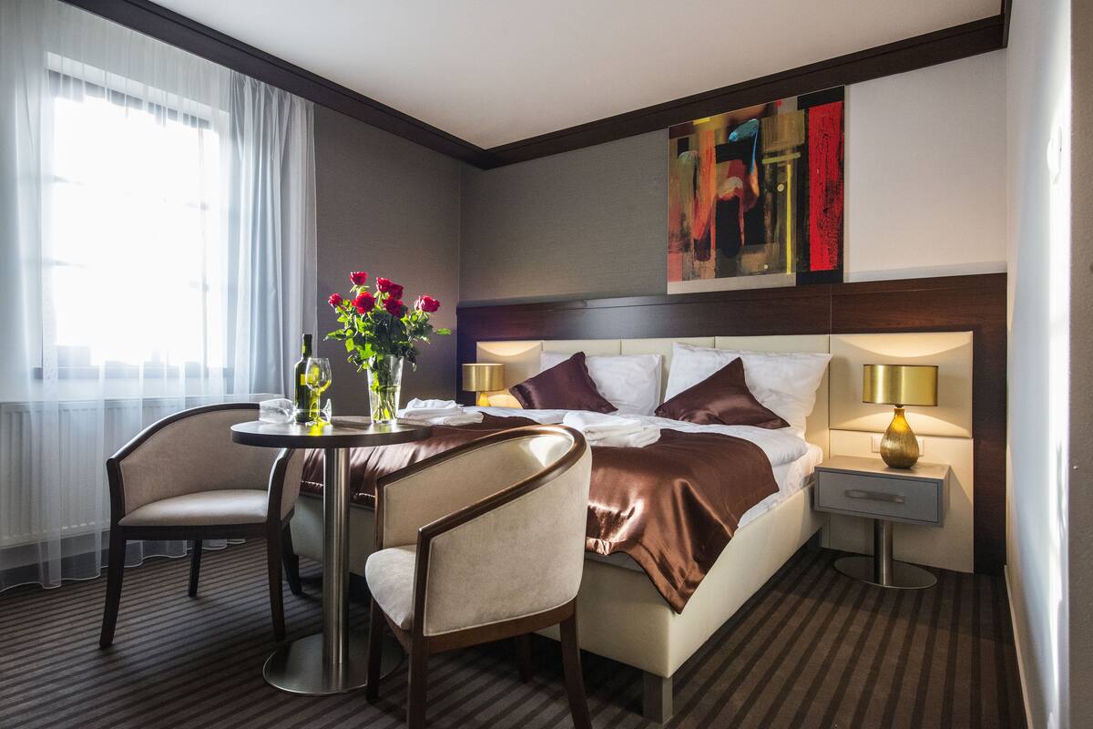 A hotel room with a bed, bedside tables, a chair, a small round table with a vase of flowers, and a window with curtains.