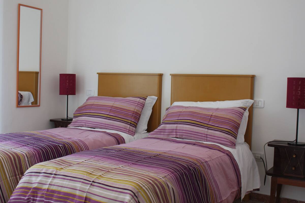 A double room with two beds, each with striped bedding and wooden headboards. There are bedside tables with lamps and a mirror on the wall.