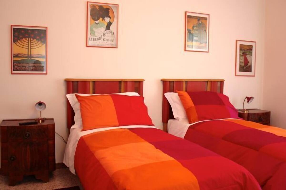 A bedroom with two beds, each with a red and orange bedspread. There are framed pictures on the wall, a bedside table with a lamp, and a small wooden cabinet.