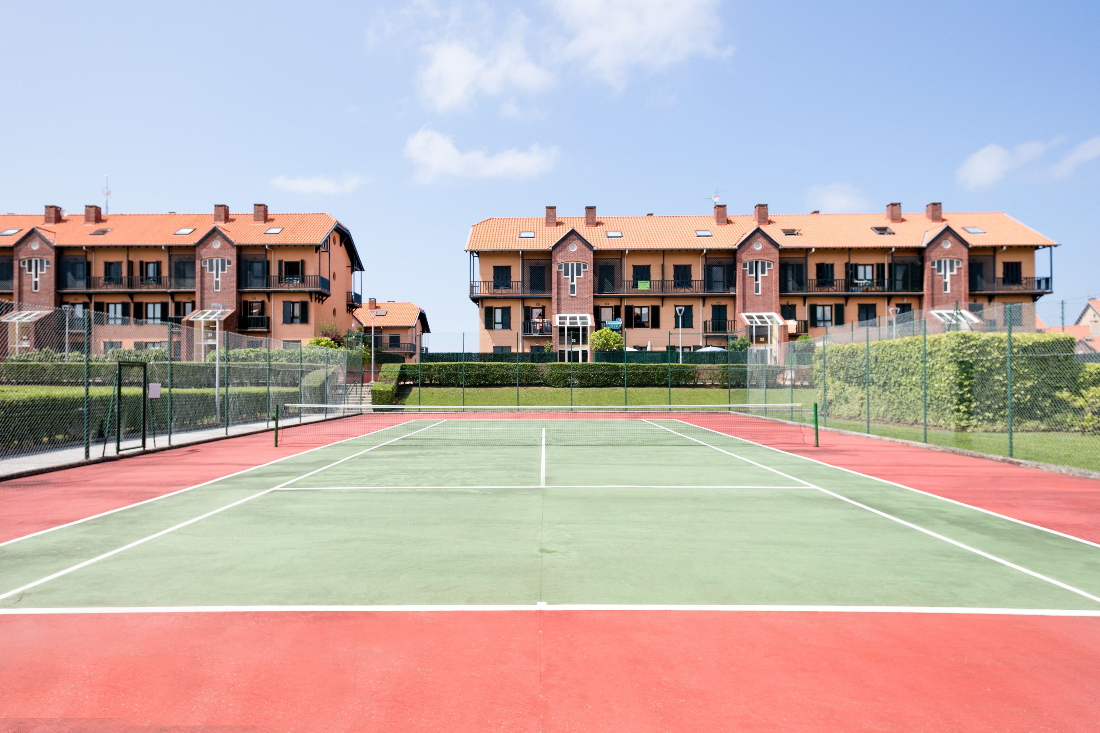 Una cancha de tenis con red en el centro, rodeada por una cerca. Se observan dos edificios con balcones al fondo.
