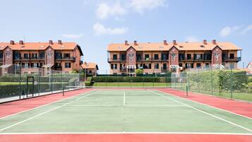 Una cancha de tenis con red en el centro, rodeada por una cerca. Se observan dos edificios con balcones al fondo.