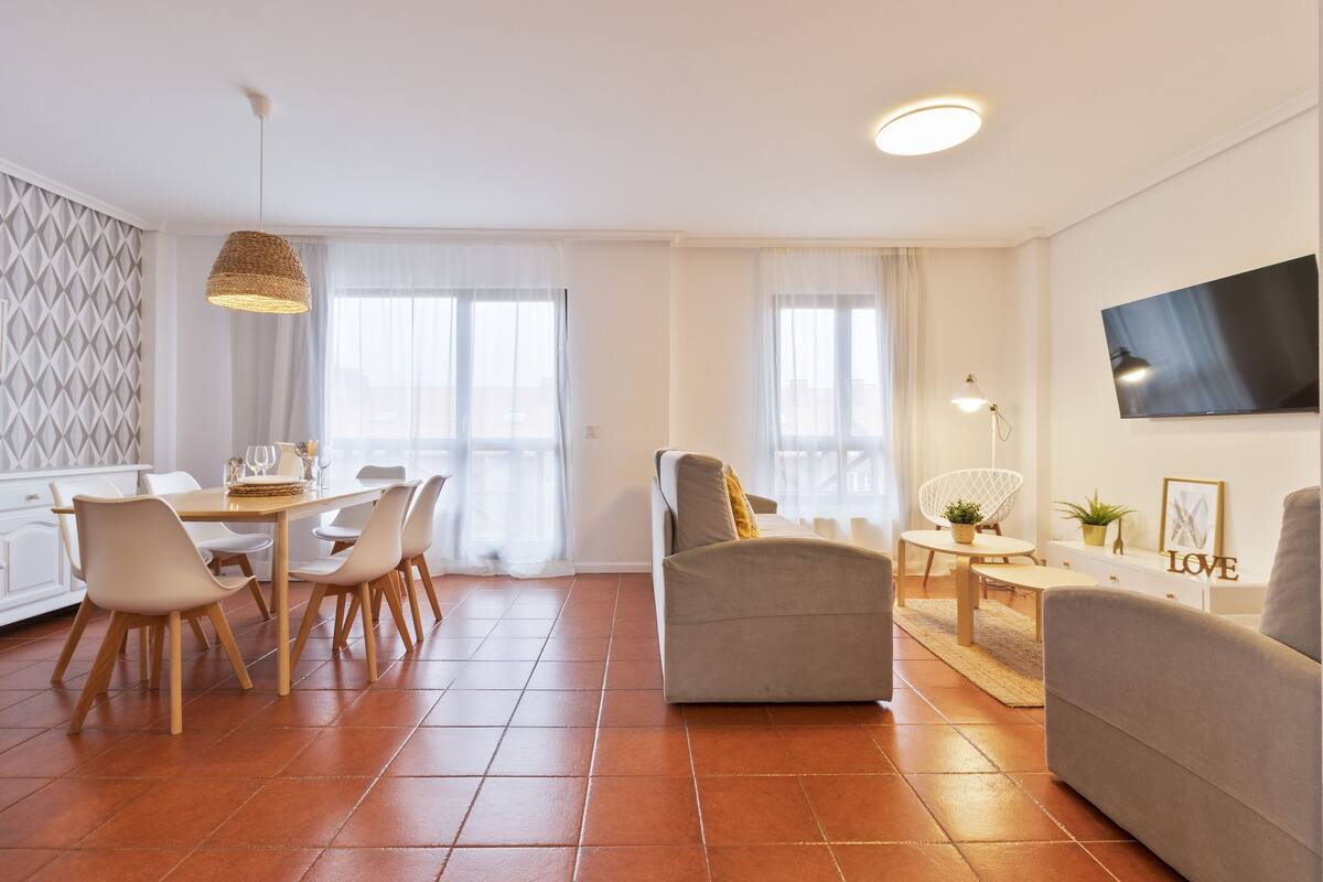 Una sala de estar moderna con un sofá, un comedor con sillas, un televisor montado en la pared y una cocina con mesa.
