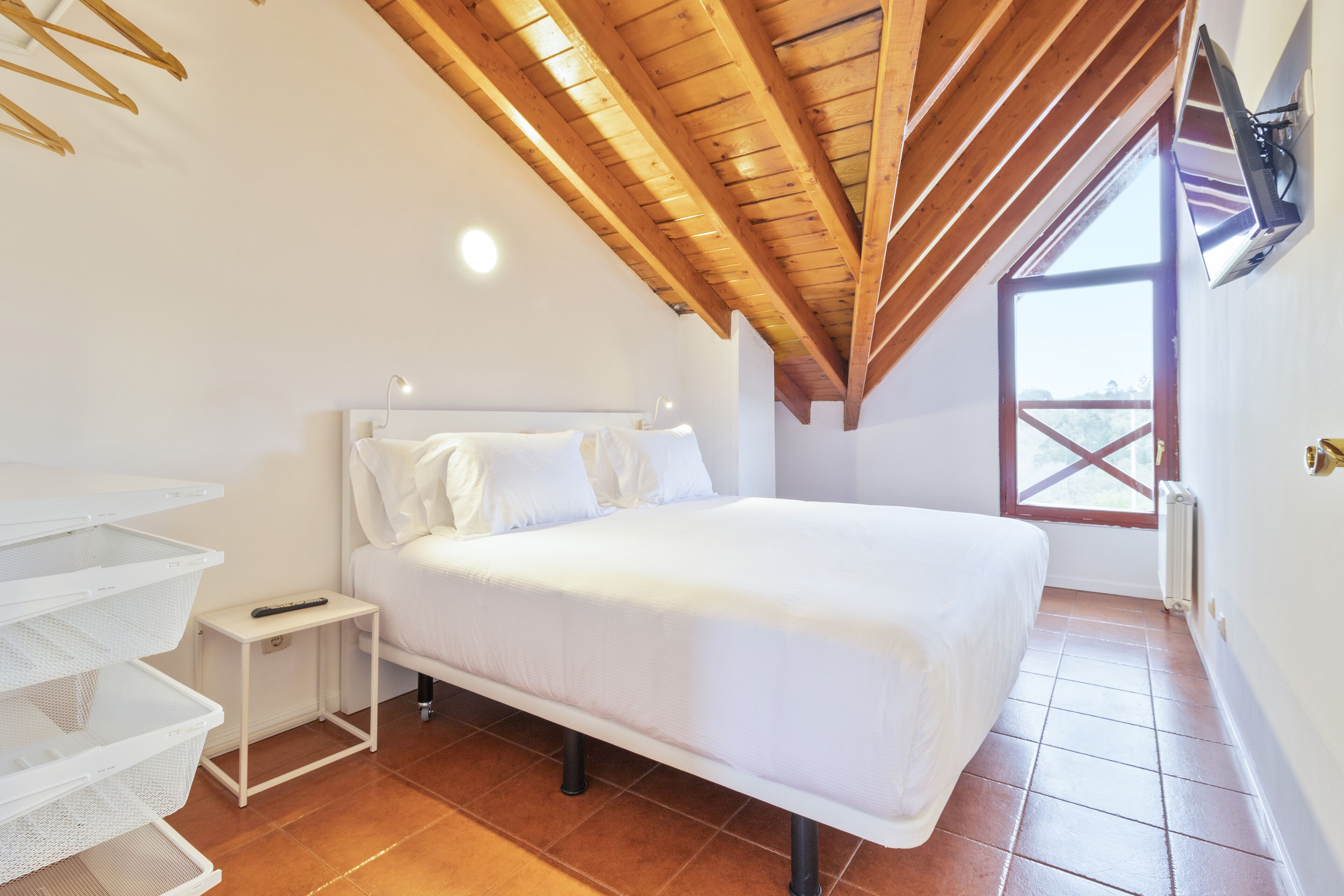 A bedroom with a wooden ceiling, a bed, white bedding, a small bedside table, and a window with a view.
