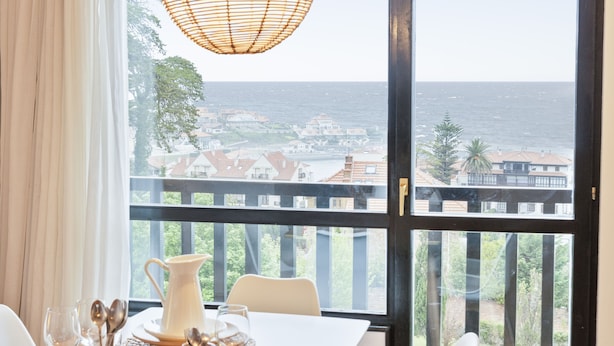 A dining table with a view of the ocean and a cityscape through large windows. - Abba Comillas Apartments (Comillas)