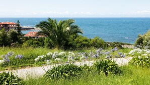 A coastal landscape with a view of the sea, lush greenery, and a pathway leading to a building with a red roof. - Abba Comillas Apartments (Comillas)