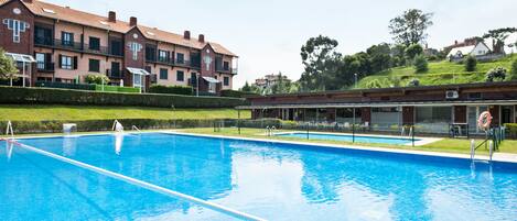 A swimming pool with clear blue water, surrounded by a well-maintained hedge and a row of brick buildings in the background.