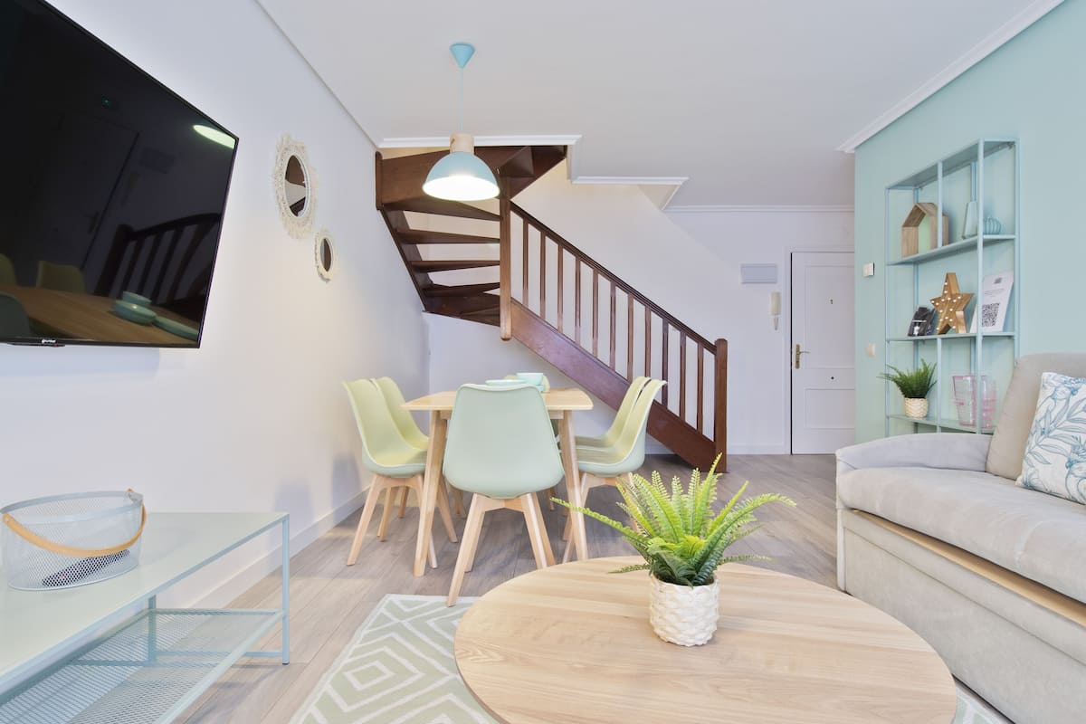 Una sala de estar moderna con una escalera, un comedor con sillas, un sofá y un televisor montado en la pared.