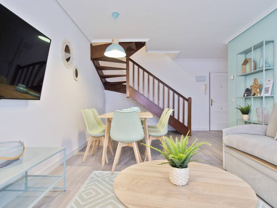 A modern living room with a staircase, a dining area with chairs, a sofa, and a TV mounted on the wall.