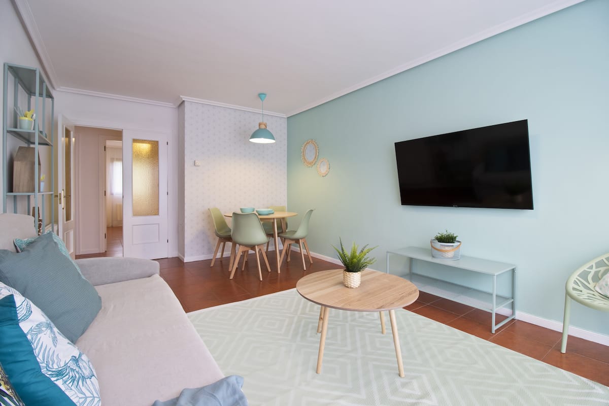 Una sala de estar moderna con un sofá, una mesa de comedor con sillas, un televisor montado en la pared y una pequeña mesita con una planta.
