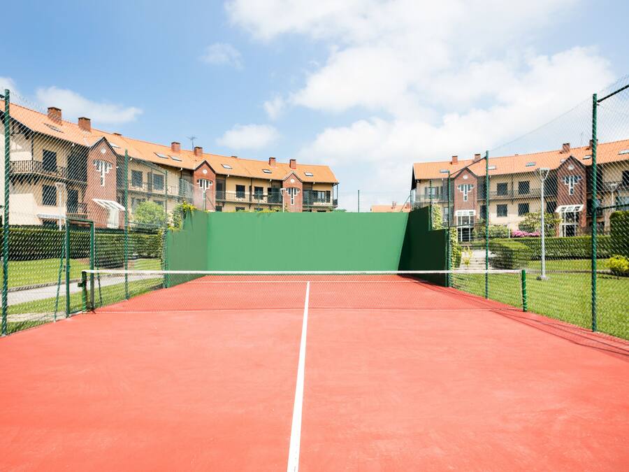 Uma quadra de tênis com um fundo verde, rodeada por edifícios residenciais com telhados vermelhos.