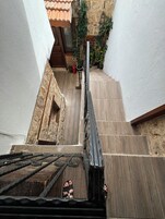 Una escalera estrecha que lleva a una puerta de madera con barandilla metálica, rodeada de paredes blancas y un muro de piedra con plantas en macetas.