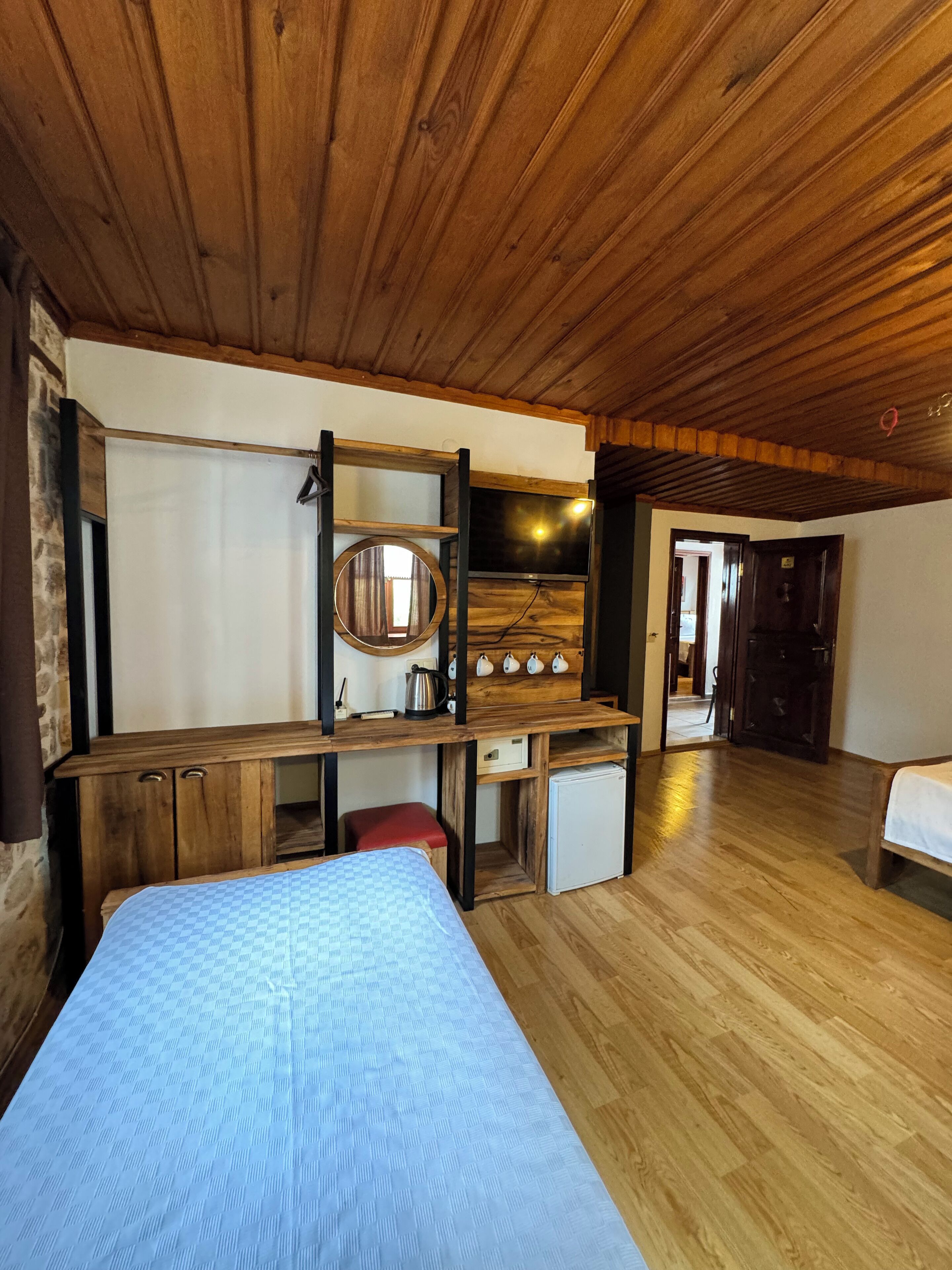 A wooden ceiling, a bed, a desk with a mirror, a small fridge, and a door leading to another room.