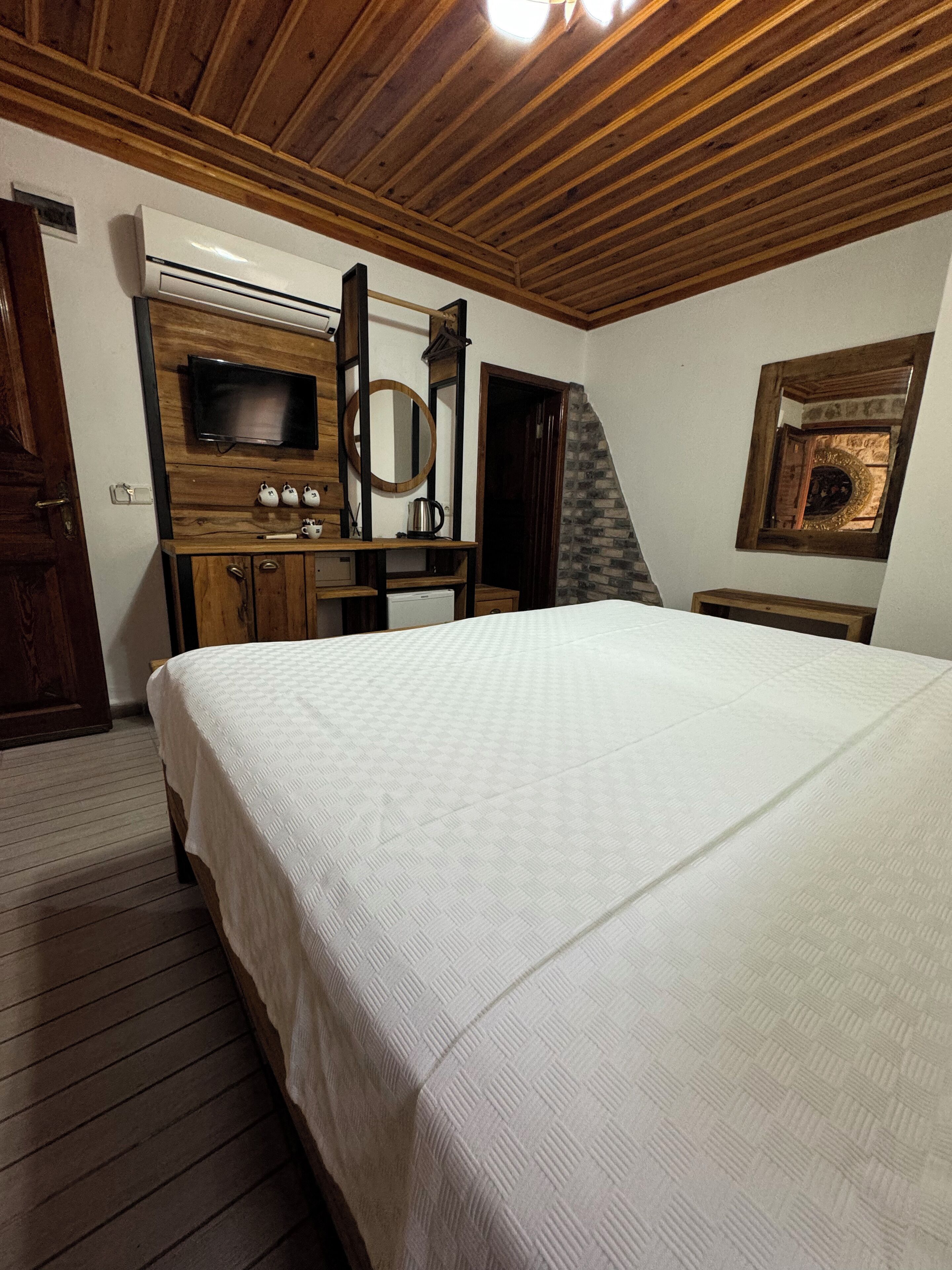 A bedroom with a large bed, wooden ceiling, a television, a mirror, and a small desk with a kettle.