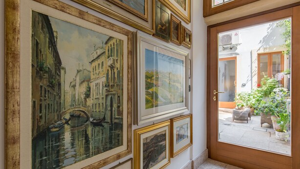 Un pasillo con cuadros enmarcados en la pared, una puerta de madera y vista a un patio exterior con plantas y una mesa.
