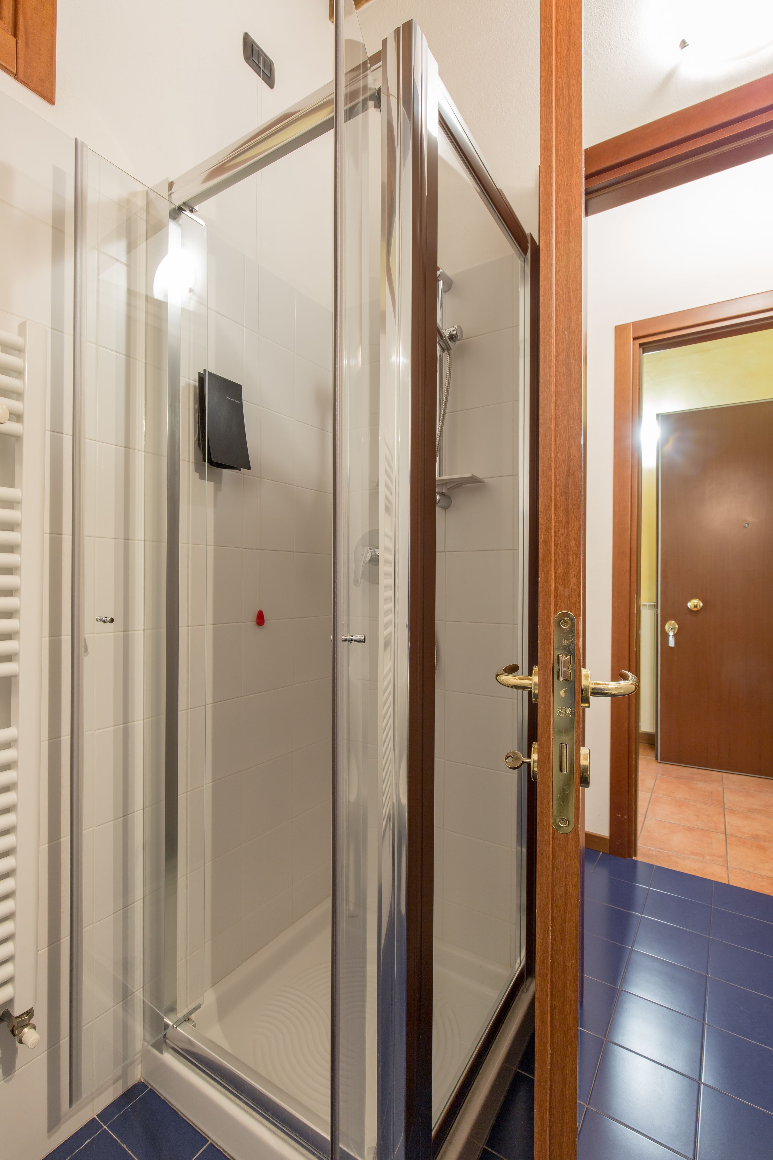 Ducha con cerramiento de vidrio, puerta de madera, paredes de azulejos blancos y piso de azulejos azules.