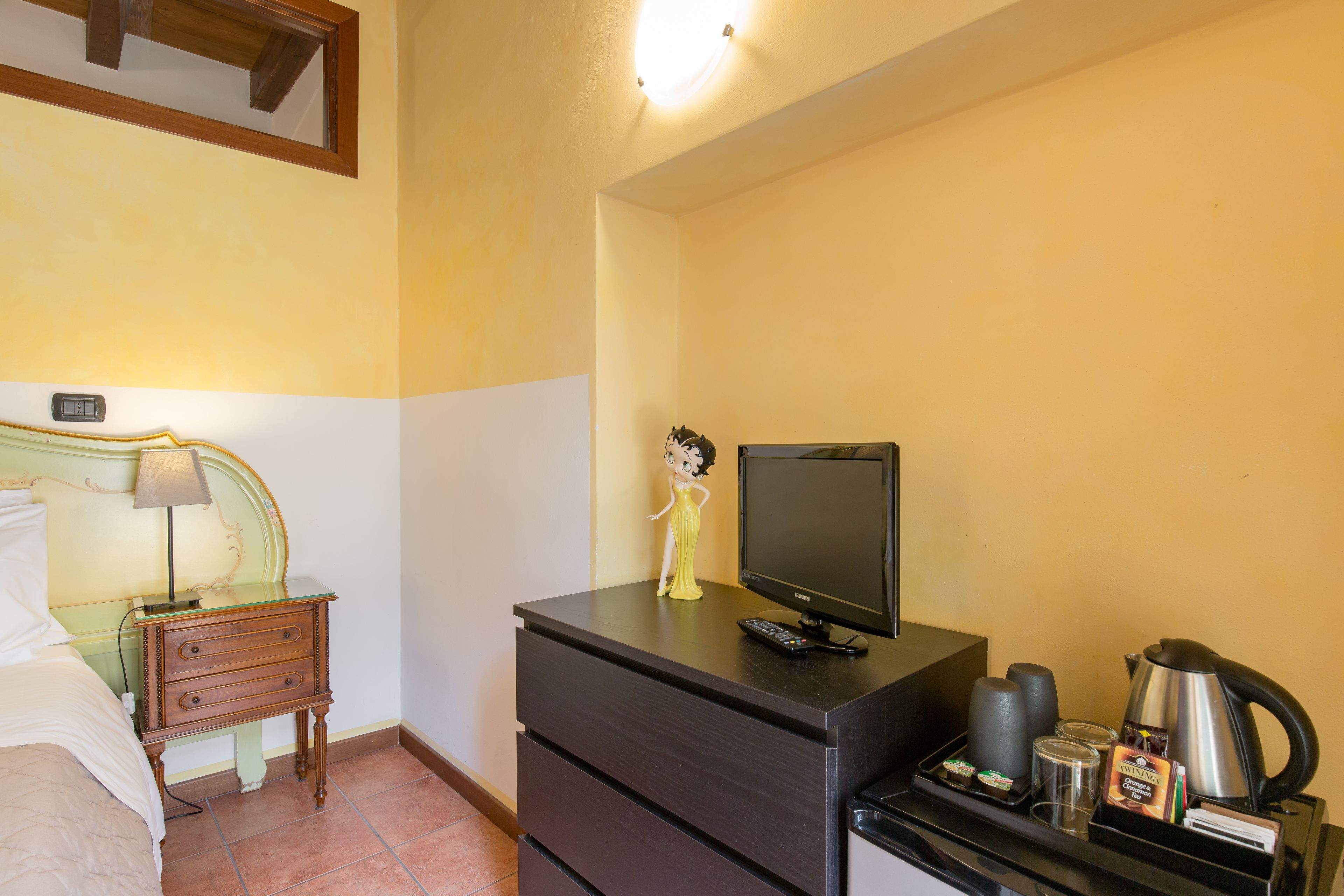 Una camera d'albergo con un letto, un comodino con una lampada, un armadio con la televisione e un vassoio con caffè e snack.