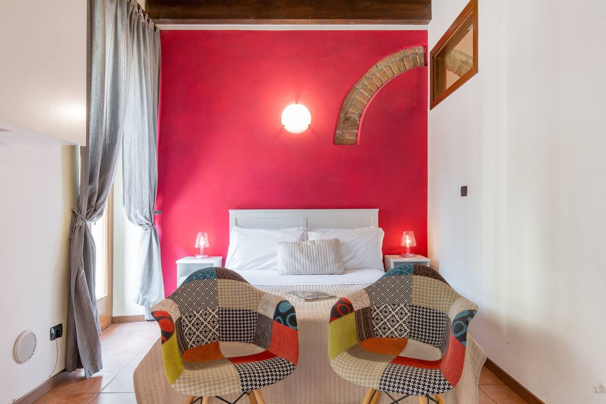A bedroom with a red wall, a white bed, two bedside tables with lamps, and two chairs with patterned upholstery.
