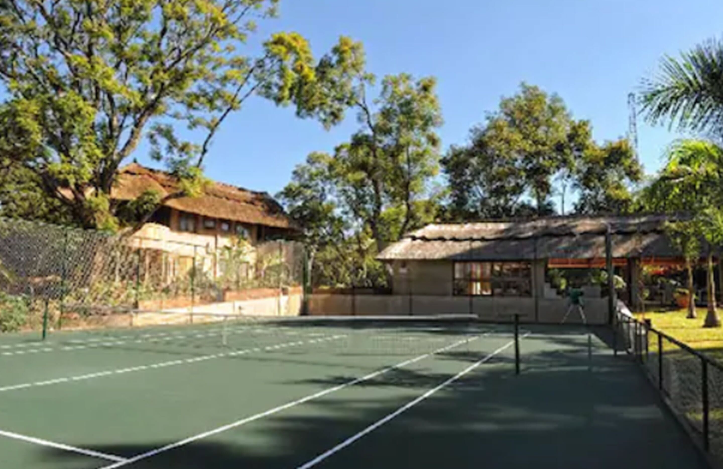 An outdoor tennis court with a net, surrounded by trees and a building with a thatched roof.
