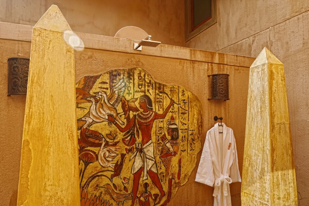 A bathroom with a mural of ancient Egyptian figures, a white robe hanging, and a tall pillar.